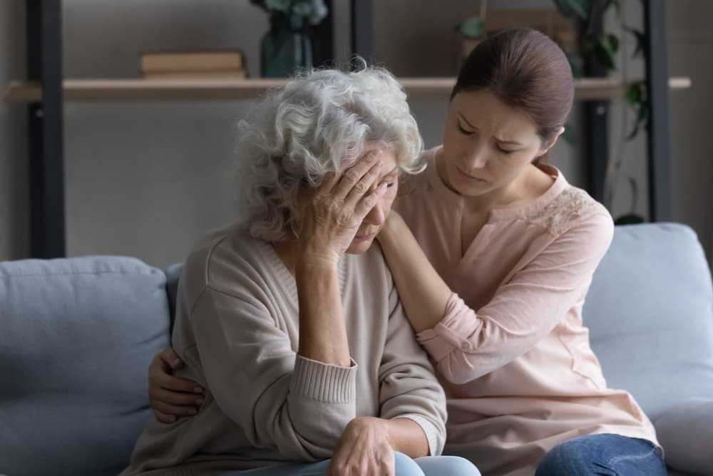 Young woman comforting upset older woman on couch at home, showing care and support, elderly female experiencing headache or discomfort.